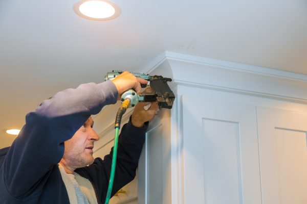 Air nailer tool close up of carpenter using nail gun to crown moldings on kitchen cabinets with white cabinets
